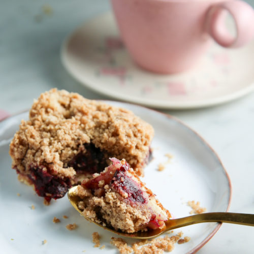 A piece of crumbly plum streusel dessert with a golden spoon rests on a white plate. Part of the dessert is scooped out, revealing a dark berry filling. In the background, a pink cup with tea sits on a matching saucer on a light-colored surface. Some crumbs are scattered around the plate.