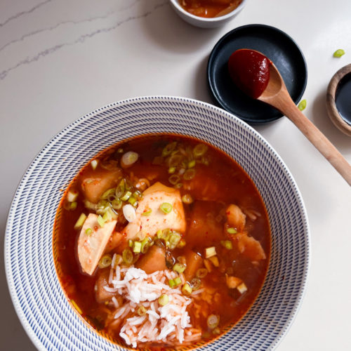 A bowl of vibrant red kimchi stew with tofu cubes, rice, and green onions is placed on a white surface. Beside the bowl are two small dishes: one with a piece of kimchi and the other with a dollop of red paste on a wooden spoon resting on the edge.