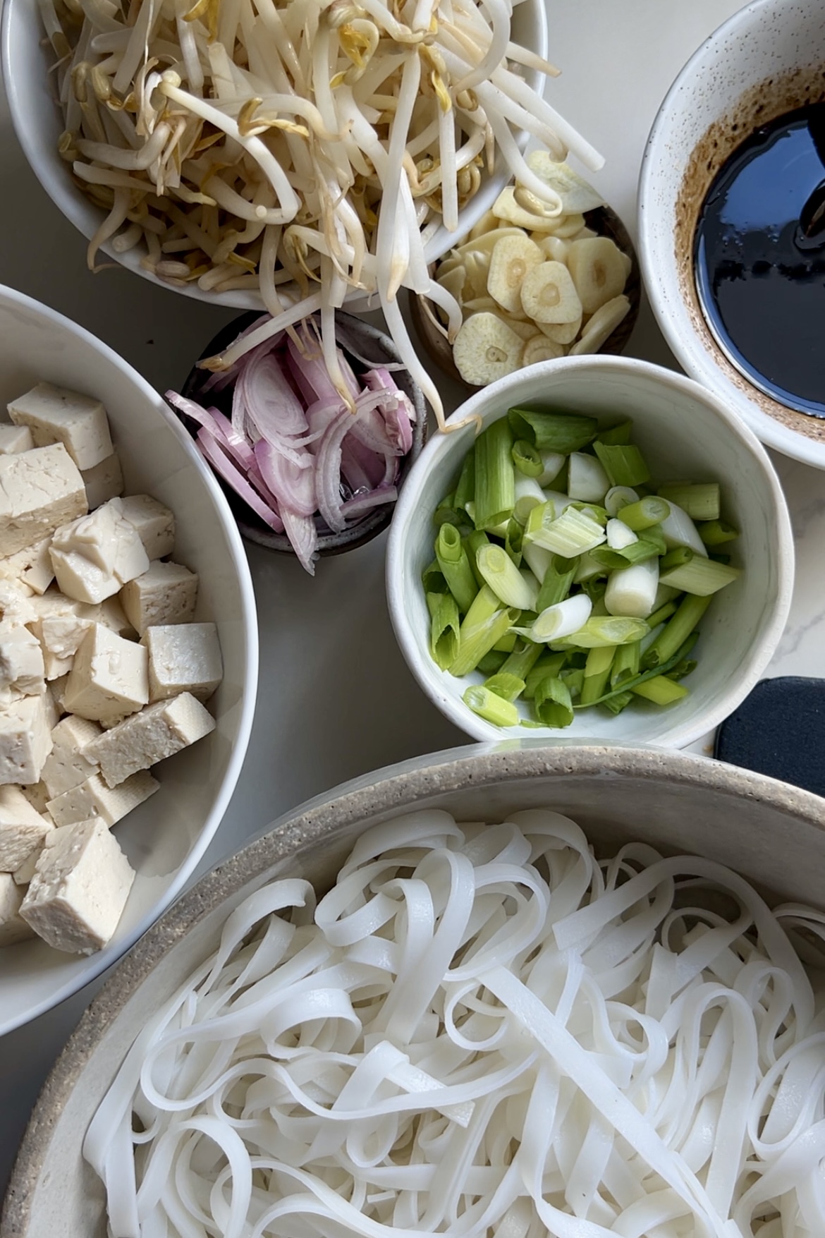 A top view of various ingredients for a meal. There are bowls containing bean sprouts, sliced garlic, sliced shallots, chopped green onions, tofu cubes, rice noodles, and a dark sauce. The ingredients are neatly arranged on a white surface, ready for cooking an authentic Pad Thai recipe.