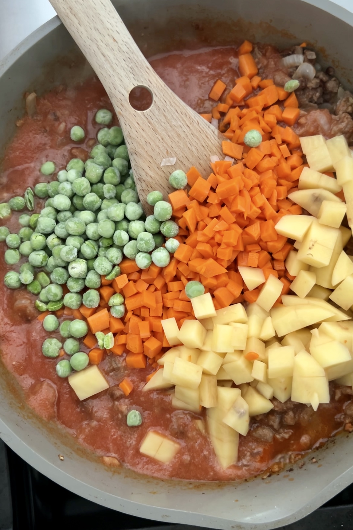 A skillet filled with a colorful mixture for cooking. It contains green peas, diced orange carrots, and yellow potatoes added to a red simmering sauce with ground meat, creating a delicious Beef Picadillo. A wooden spoon rests in the pan, partially submerged in the ingredients.