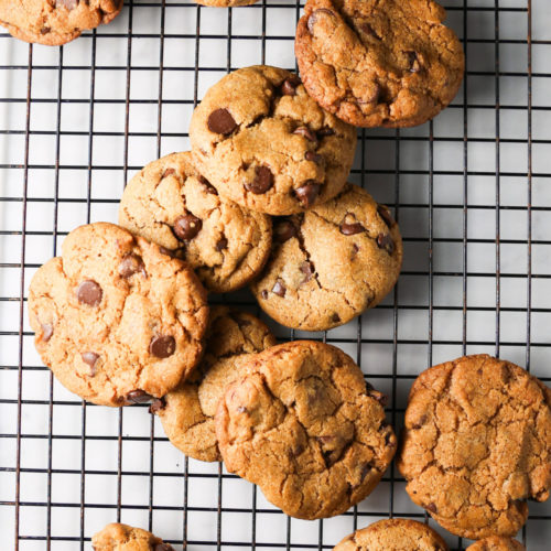 A batch of chocolate chip cookies is cooling on a black wire rack. The golden brown cookies, infused with a hint of pumpkin spice, have visible chocolate chips and slightly cracked surfaces. One cookie is broken in half, and the overall arrangement is casual, with some cookies overlapping unevenly on the rack.