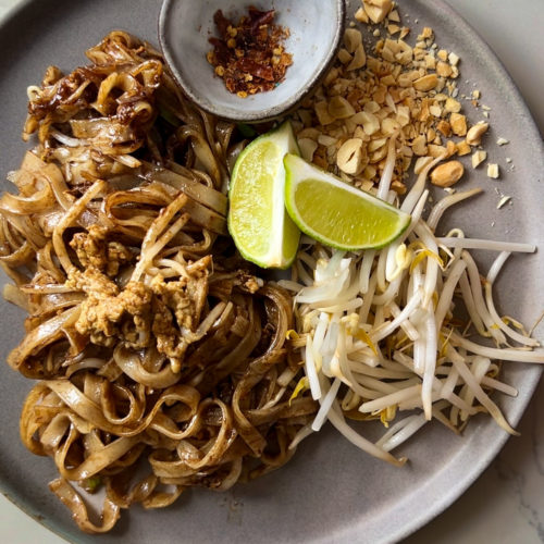 A plate of Pad Thai featuring stir-fried noodles with egg, tofu, and seasonings is garnished with bean sprouts, crushed peanuts, two lime wedges, and a small dish of red chili flakes. The meal is presented on a gray plate on a light-colored surface.