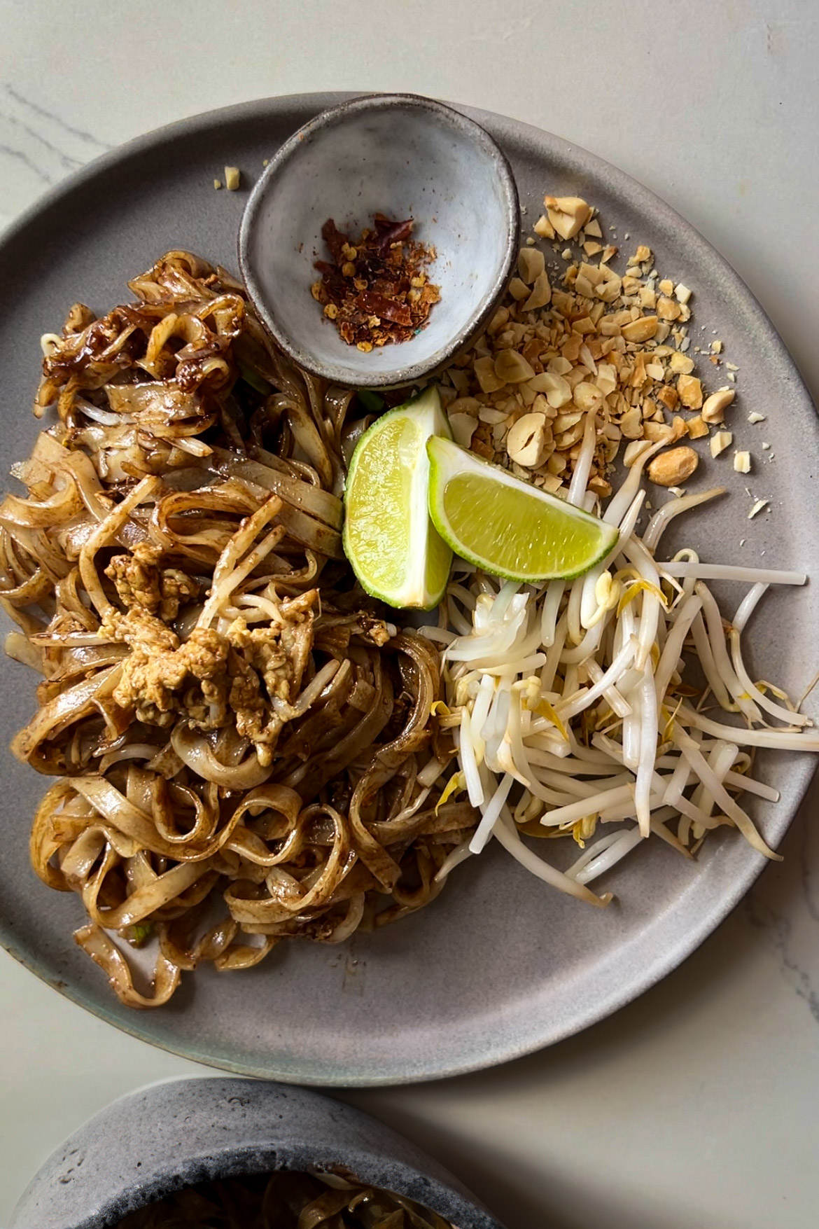 A plate of Pad Thai featuring stir-fried noodles with egg, tofu, and seasonings is garnished with bean sprouts, crushed peanuts, two lime wedges, and a small dish of red chili flakes. The meal is presented on a gray plate on a light-colored surface.