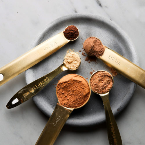 A set of five brass measuring spoons, akin to an artist’s auto draft, is elegantly filled with various spices on a round, gray plate. Labeled precisely from 1/4 tsp to 1 tbsp, the spices present a palette of browns and reds against a marble background.