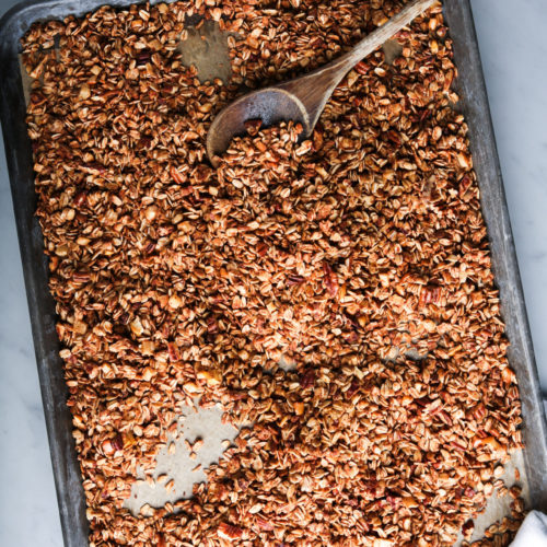 A large baking sheet filled with baked granola sits on a marble counter. The granola is golden brown and textured, with visible oats and nuts. A wooden spoon rests on the granola, suggesting it has been recently stirred. In the corner, a white cloth is slightly visible.