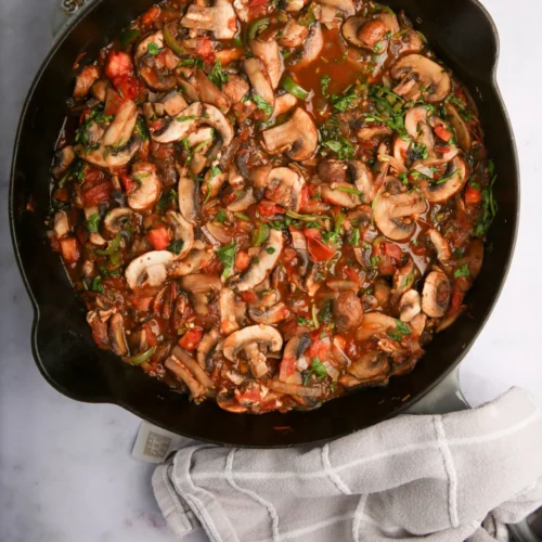 A black skillet filled with Champiñones a la Mexicana, garnished with fresh herbs. The sliced mushrooms mingle with diced tomatoes for a colorful, hearty appearance. A gray checkered kitchen towel sits beside the skillet on the marble countertop.