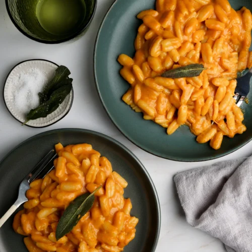 Dos platos verdes están llenos de pasta de calabaza fácil en una salsa cremosa de naranja, adornada con hojas de salvia. Un tenedor con mango negro reposa sobre cada plato. Cerca hay un vaso verde con un líquido misterioso, un pequeño cuenco de sal y una servilleta de tela beige, todo sobre la superficie de mármol claro.