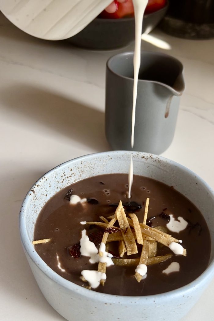 A light blue bowl of Mexican bean soup, also known as crema de frijol, is being garnished with a stream of cream from a gray pitcher in the background. The soup is thick and dark with pieces of tortilla strips and what appears to be dried chili pepper bits on top. The setting is on a light-colored countertop.