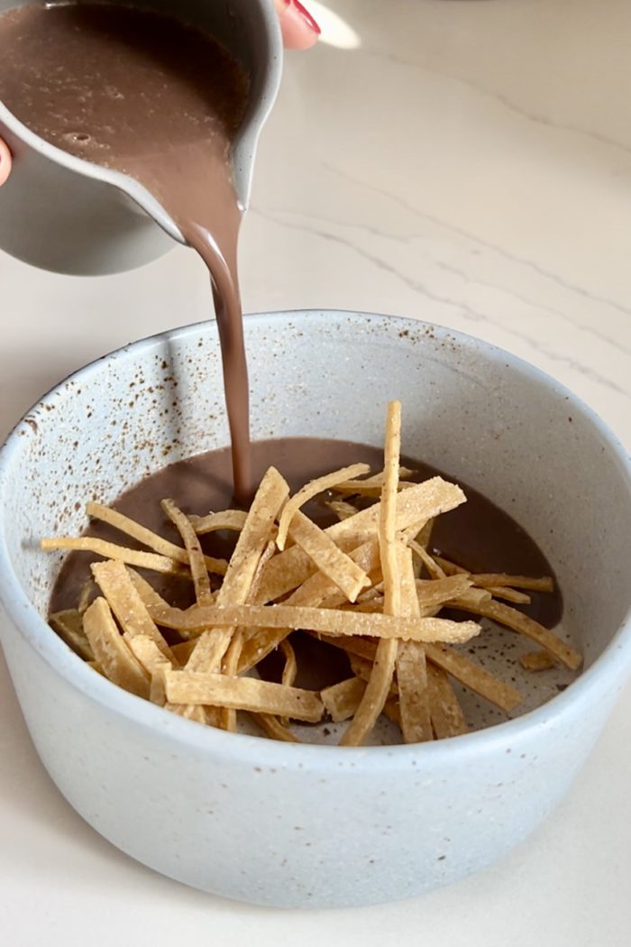 A hand pours a thick, brown sauce from a small pitcher into a light blue speckled bowl filled with crispy, golden tortilla strips. The bowl sits on a light-colored countertop. The scene implies the preparation of Crema de frijol, potentially creating a delicious Mexican creamy bean soup.