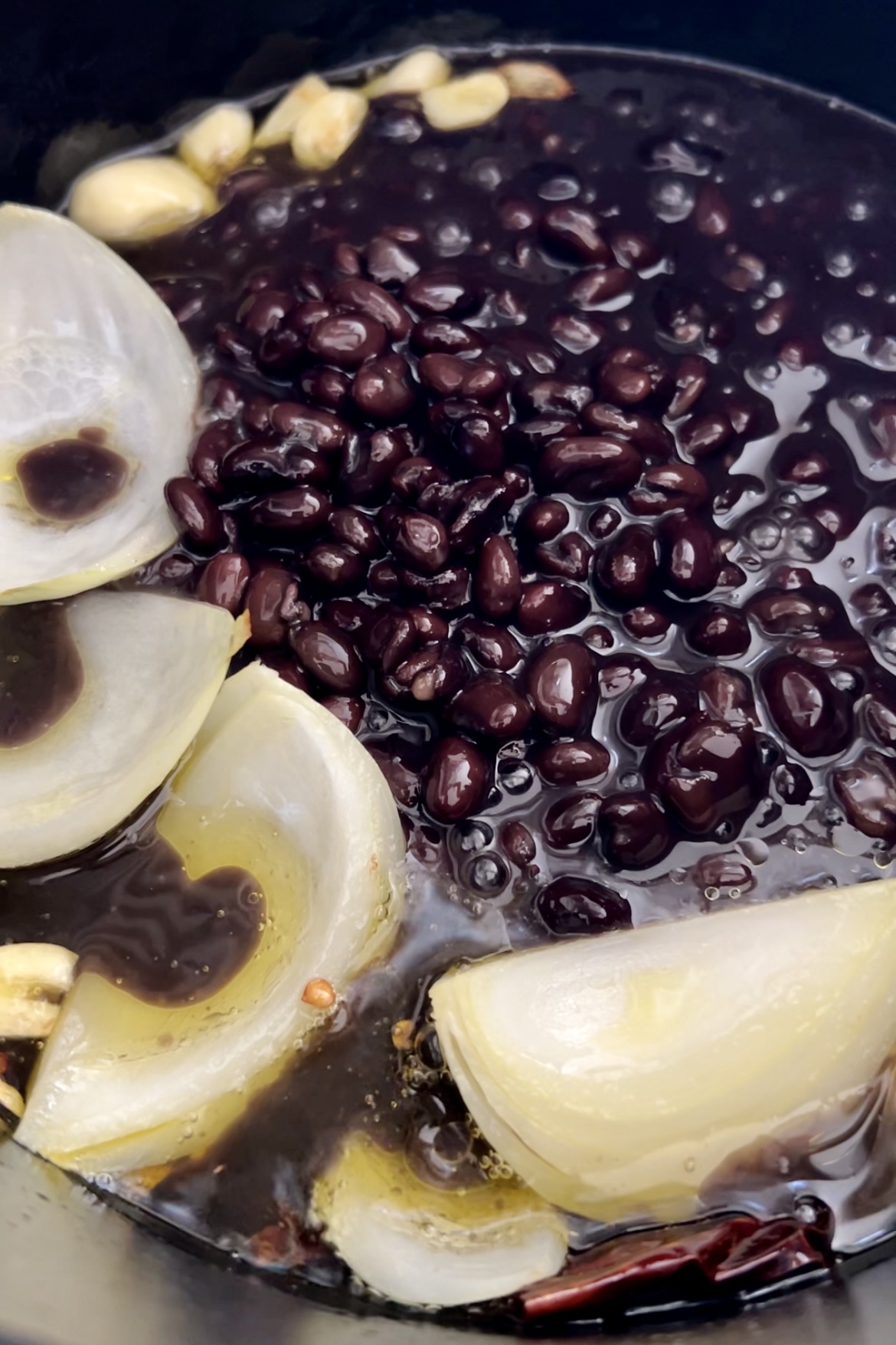 Black beans simmer with large onion pieces, garlic cloves, and red chili peppers in a dark, flavorful broth. The various ingredients float amidst the rich liquid in a cooking vessel, suggesting the early stages of preparing Crema de frijol, a hearty Mexican bean soup.