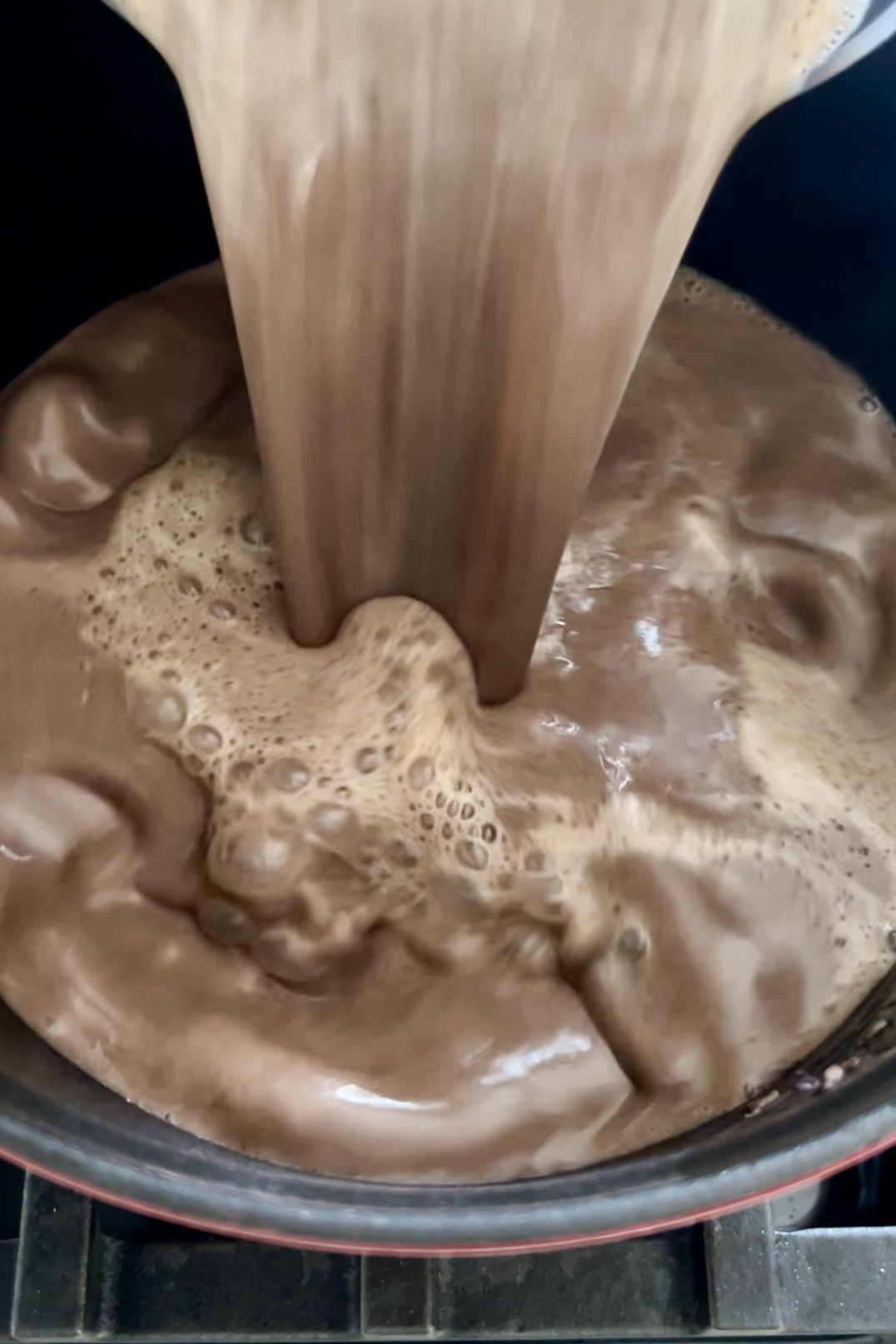 Close-up of a thick, creamy liquid being poured into a pot or container. The liquid appears chocolatey, creating bubbles and froth on the surface as it splashes and swirls. The image conveys the rich, velvety consistency of crema de frijol, highlighting its motion and texture.