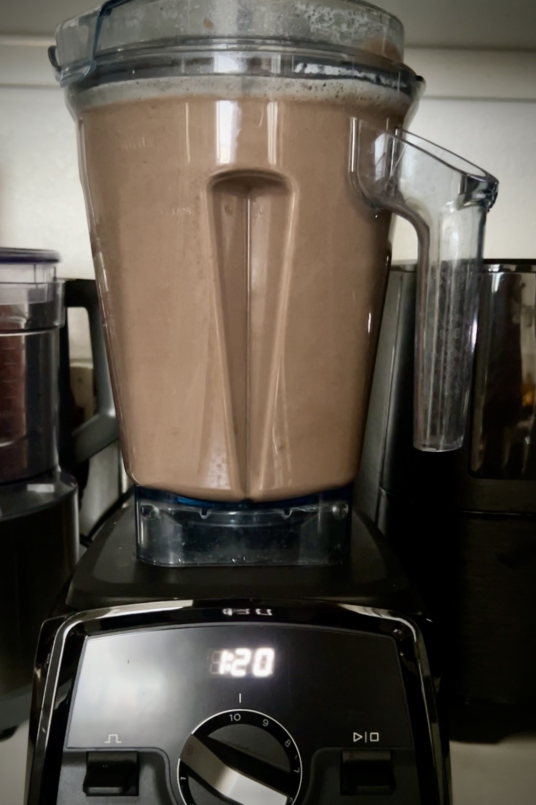 A blender filled with a brown crema de frijol smoothie is placed on a kitchen counter. The digital display on the blender base indicates the number '1:20'. Other kitchen appliances are visible in the background, such as a dark-colored coffee grinder on the left and an air fryer on the right.