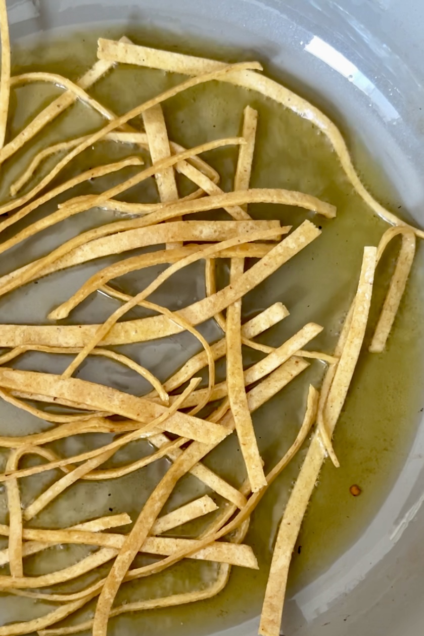Thinly sliced tortilla strips are being lightly pan-fried in oil in a gray skillet. The strips are golden-brown and irregularly arranged. The oil appears hot, with a few small bubbles visible around the edges of the tortilla strips—perfect toppings for Crema de Frijol.