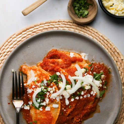 A gray plate on a woven mat showcases Mexican cuisine at its finest: entomatadas drenched in red sauce, garnished with crumbled cheese, sliced onions, and chopped cilantro. A fork lies beside them. Above, a small bowl of sauce with a spoon sits alongside wooden bowls of fresh cilantro and cheese.