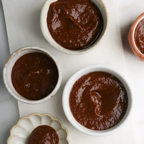 Four small bowls filled with rich, glossy chocolate ganache are arranged on a white marble surface. A wooden spoon rests on a small flower-shaped dish, holding a dollop of the homemade ganache. The setup creates a visually appealing presentation of this delightful dessert.