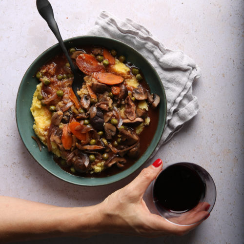 A hand with red nail polish holds a glass of red wine next to a green plate of polenta topped with mushroom bourguignon containing carrots, peas, and sauce. A black spoon rests in the bowl, which is placed on a light gray table with a textured white cloth nearby.