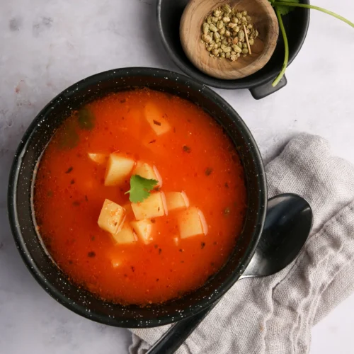 A black bowl cradles a vibrant red-orange Mexican Potato Soup dotted with cubes of potato and garnished with cilantro. Next to it, a small black dish holds coriander seeds and a cilantro sprig. Nearby, on the white marble surface, rest a gray napkin and a black spoon.