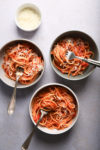 Three bowls of spaghetti with tomato sauce and grated cheese, each with a fork, are arranged on a light gray surface. A small bowl of extra grated cheese is placed above the bowls. The image has a top-down perspective, showcasing the vibrant colors and textures.