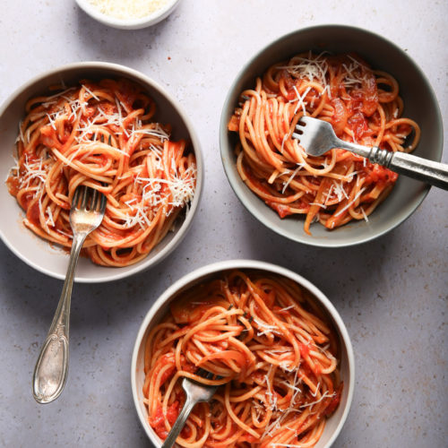 On a light surface, three bowls of spaghetti with tomato sauce and grated cheese are arranged. Each bowl contains a fork. Above the bowls, there is a small plate with extra grated cheese, ready to serve. The well-lit scene highlights the textures of the pasta, and the fennel seeds add an aromatic touch.