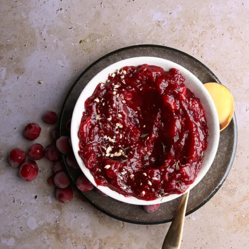 A bowl of the easiest-to-make cranberry sauce, garnished with chopped nuts, rests on a dark plate with a gold spoon. Fresh cranberries scattered on the textured surface add a rustic touch to this charming composition.