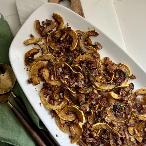 A white rectangular plate showcases roasted delicata squash with a savory pecan crumble, all highlighted by a maple chipotle glaze. The dish rests on a wooden and marble board. Beside it, a green cloth napkin and gold utensils with wooden handles sit elegantly on the white countertop.