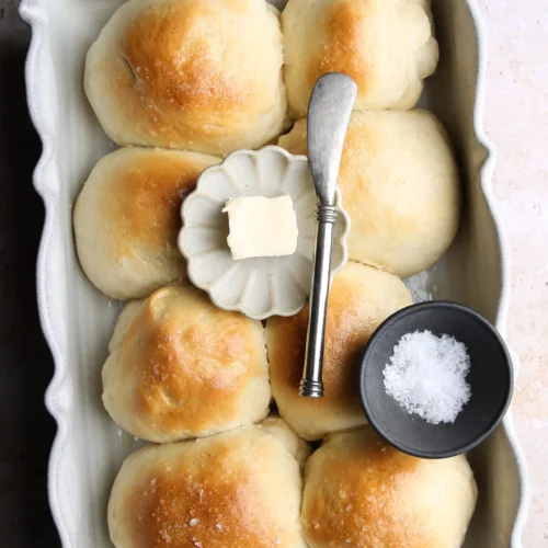 Six golden-brown dinner rolls are nestled in a rectangular white ceramic dish. A small plate with a pat of butter and a butter knife rests on top, ready to enhance the flavor. A small black bowl of salt lies nearby, echoing the light dusting atop the rolls. The marbled beige surface adds a touch of elegance.