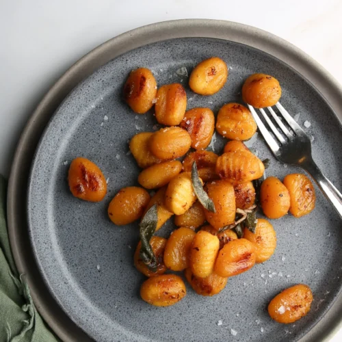 Top-down view of a gray plate with pan-seared golden brown gnocchi drizzled with lemon brown butter sauce, garnished with crispy sage leaves and sprinkled with sea salt. A fork rests on the plate. The background is a white marble surface, and a green cloth napkin is partially visible on the side.