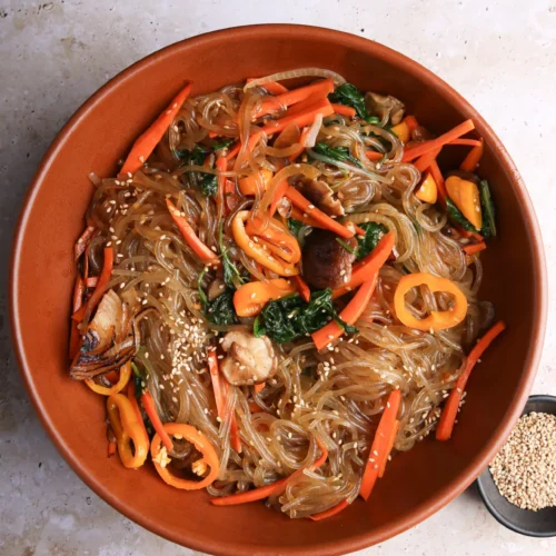 A large orange bowl of delicious Japchae, featuring stir-fried Korean glass noodles intertwined with sliced carrots, vibrant bell peppers, shiitake mushrooms, spinach, and sesame seeds. The backdrop is a textured light stone surface with a small black dish of sesame seeds nearby.