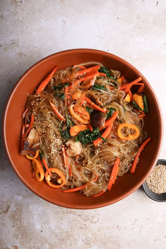 A large orange bowl of delicious Japchae, featuring stir-fried Korean glass noodles intertwined with sliced carrots, vibrant bell peppers, shiitake mushrooms, spinach, and sesame seeds. The backdrop is a textured light stone surface with a small black dish of sesame seeds nearby.