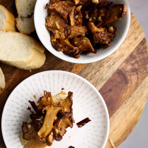 Two white bowls filled with garlicky mushrooms rest on a wooden board. The larger bowl features mushrooms garnished with herbs, while the smaller plate holds a baguette slice topped with mushrooms. Additional bread slices are beside the bowls, set against a light gray background.