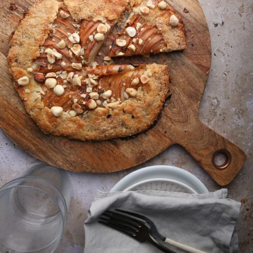 A rustic pear galette topped with chopped hazelnuts and a hint of blue cheese sits on a round wooden board. One slice is slightly pulled away. Beneath, a marble surface holds a glass of water and a white, textured plate with a grey napkin, fork, and knife resting on it.