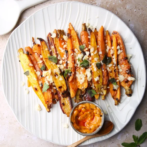 A plate of spicy roasted multicolored carrots garnished with crumbled cheese and fresh herbs. Accompanied by a small dish of creamy orange dipping sauce and a wooden spoon. The background is a textured, light-colored surface, with a white spatula partially visible in the upper left corner.