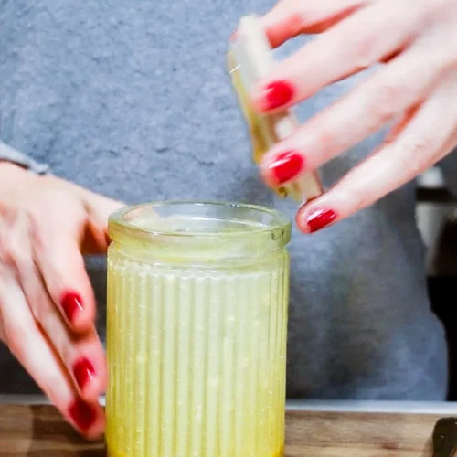 A person with red nail polish is placing the lid on a ridged glass jar filled with a yellow apple cider vinaigrette. They are wearing a gray sweater. The jar sits on a wooden surface, and a spoon is partially visible in the foreground.