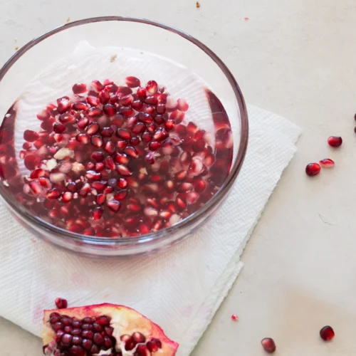 A bowl of water, brimming with pomegranate seeds, sits on a white napkin. Surrounding it are scattered seeds and a partially peeled pomegranate on a light-colored surface, creating an easily elegant scene.