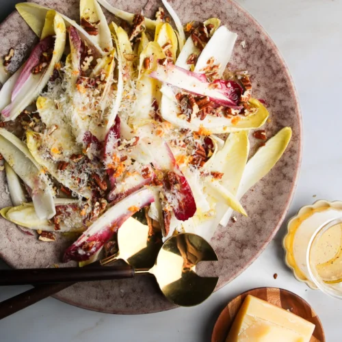 A platter of chicory salad with sliced endives, shaved cheese, chopped pecans, and a sprinkle of spices offers a taste from this delightful endive salad recipe. A wedge of cheese and a bowl of creamy dressing are nearby, with a fork and spoon resting on the plate against the light, textured background.