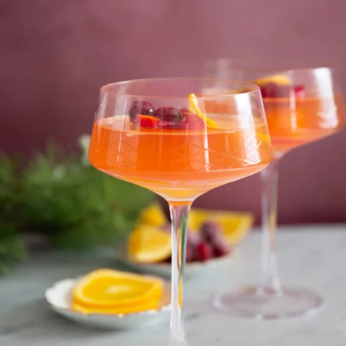 Two elegant coupe glasses filled with a vibrant, festive mimosa are garnished with cranberries and lemon twists. They rest on a marble surface with orange slices and scattered cranberries in the background. The setting features a blurred greenery backdrop, adding a touch of natural elegance.
