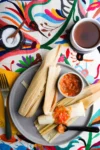 A plate of tamales drizzled with tomato salsa is on a vibrant, embroidered tablecloth. One tamale is partially unwrapped, showcasing its savory filling. A cup of tea sits to the upper right, next to a bowl of salt with a spoon. A fork rests on a yellow napkin beside the plate, hinting at an inviting meal enhanced by olive oil.