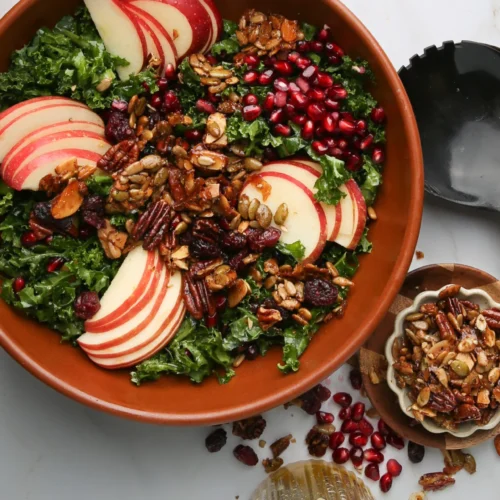 A brown bowl brimming with kale salad is topped with sliced red apples and pomegranate seeds, all enhanced by a mix of nuts. Nearby, a small bowl of nuts and a black serving spoon await. A glass of apple cider vinaigrette sits below, surrounded by scattered seeds on the white surface.
