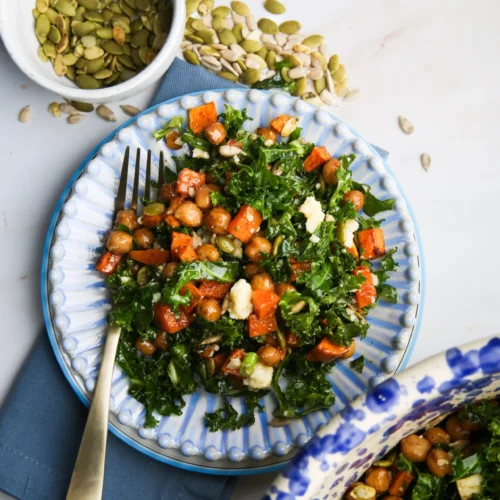 A colorful kale salad with roasted sweet potatoes, chickpeas, and crumbled feta is drizzled with maple Dijon dressing and served on a decorative blue plate. A fork rests on the plate atop a blue napkin, while small bowls alongside hold pumpkin and sunflower seeds. A large bowl with more salad is partially visible.