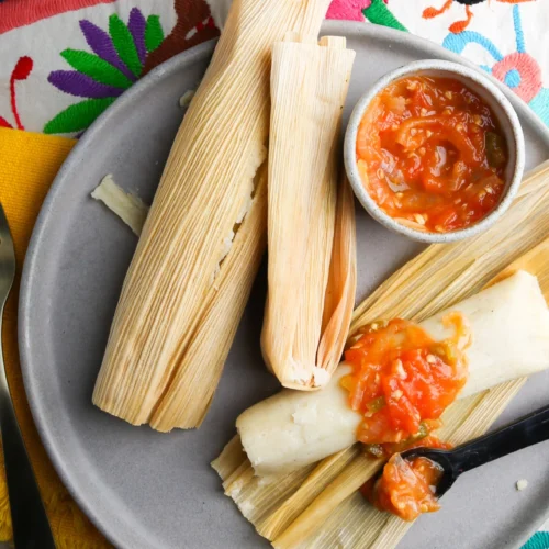 A plate featuring two tamales, one partially unwrapped and topped with red salsa enriched with a hint of olive oil. The tamales are wrapped in corn husks, and a small bowl of salsa sits beside them. The vibrant background of colorful embroidered fabric highlights the cozy scene. Cups peek into view.