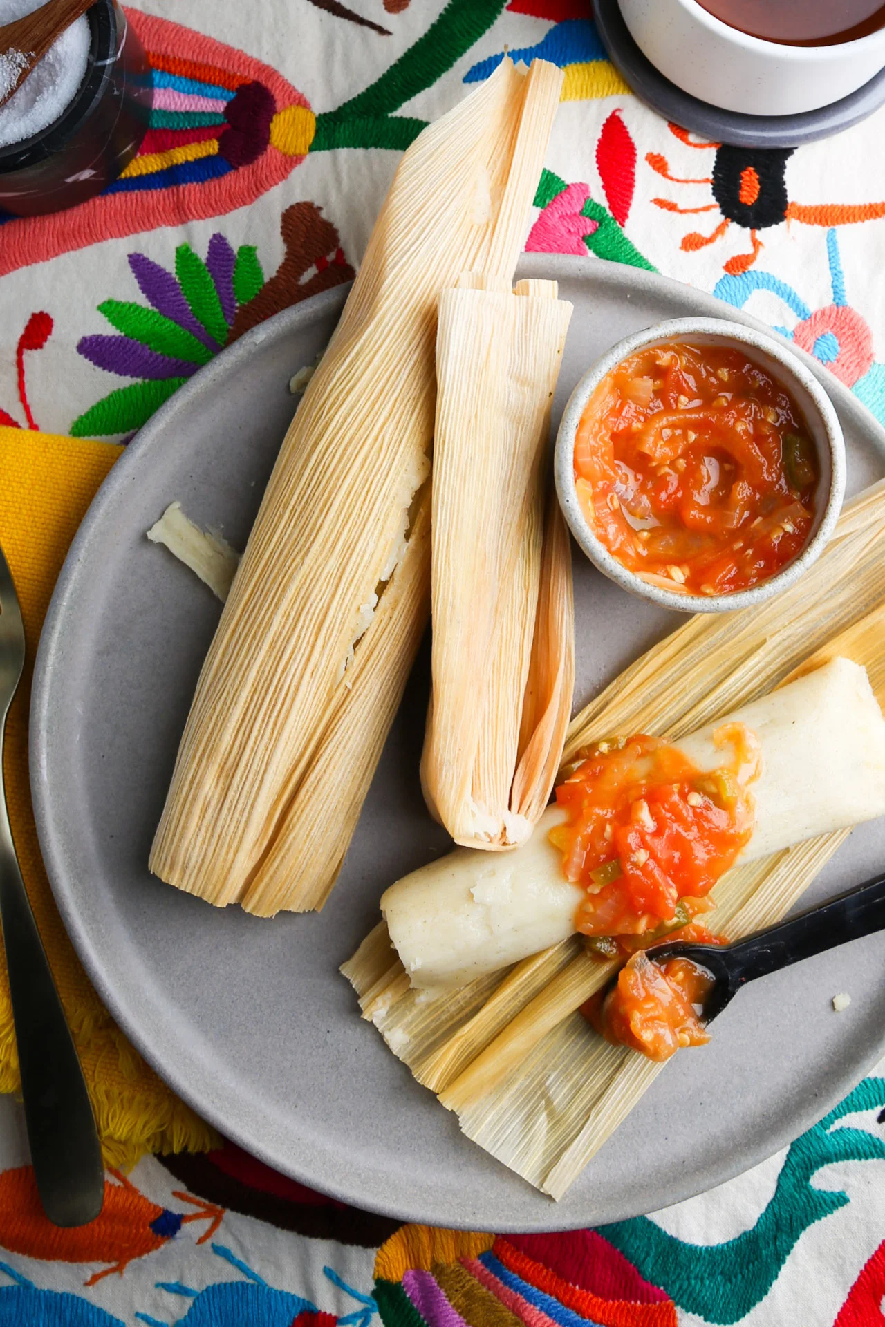 A plate featuring two tamales, one partially unwrapped and topped with red salsa enriched with a hint of olive oil. The tamales are wrapped in corn husks, and a small bowl of salsa sits beside them. The vibrant background of colorful embroidered fabric highlights the cozy scene. Cups peek into view.