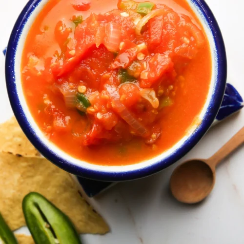 A ceramic bowl filled with vibrant red salsa, featuring diced tomatoes, onions, and jalapeños. The bowl is on a white surface with tortilla chips and sliced jalapeños nearby. A small wooden spoon lies to the right of the bowl.