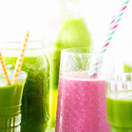 A pink smoothie in a clear glass with a blue and pink striped straw takes center stage as the ultimate smoothie. Surrounding it are various jars filled with green smoothies, each adorned with different striped straws. The bright background highlights the vibrant colors of these refreshing drinks.