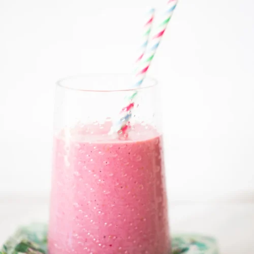 A frothy pink smoothie in a clear glass, with two colorful striped straws. The glass is placed on a stack of patterned, colorful napkins. The background is white and softly blurred, highlighting the vibrant pink of the smoothie.