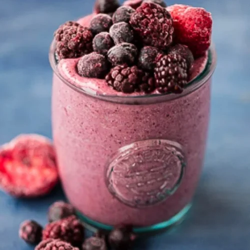 A glass filled with a thick, berry smoothie topped with a variety of frozen delights, including blackberries, raspberries, and strawberries. Scattered around the base are more berries. There's even a hint of creamy avocado blended in. The background is a textured blue surface.