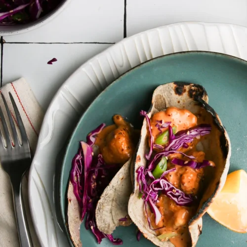 A vibrant plate with two shrimp tacos drizzled in creamy orange sauce, topped with shredded purple cabbage and green onions. A lemon wedge rests on a teal plate above a larger white one. To the left, a fork lies on a striped napkin near a bowl of cauliflower florets.