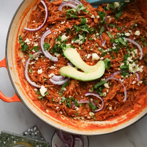 A skillet brimming with Fideo Seco, reddish-brown noodles enhanced by chipotle, is topped with avocado slices, crumbled cheese, and red onion rings. Chopped greens are sprinkled across. A blue spatula rests inside. Nearby are a drink, a small bowl of red onion, and cheese on a green napkin.