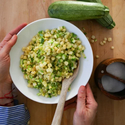 A person in a striped shirt holds a bowl of corn and chopped green vegetables, crafting a Sinaloense colache with a wooden spoon. Two zucchinis rest on the wooden table nearby, alongside a small bowl containing two gray stones.