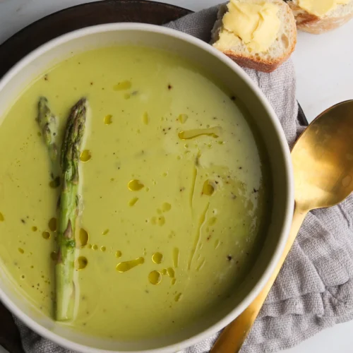 A bowl of creamy asparagus soup, made even richer with cashew milk, is garnished with whole asparagus spears and drizzled with olive oil on a wooden plate. Beside it, a gold spoon and slices of crusty bread with butter rest on a gray napkin against a smooth, light-colored surface.
