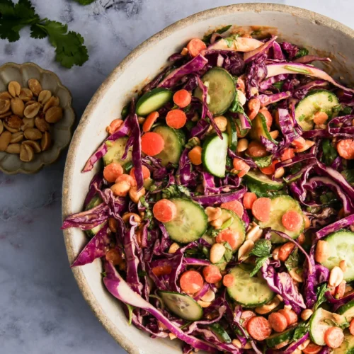 A vibrant Crunchy Red Cabbage Salad in a bowl features sliced cucumbers, carrots, red cabbage, and peanuts, all mixed with fresh cilantro. To the side, there's a small dish containing more peanuts. The light marbled surface background enhances the colorful ingredients.